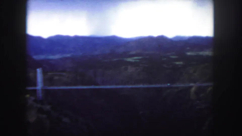 Colorado 8mm still with a suspension bridge crossing a dark canyon beneath blue-toned mountain ridges and a bright sky.