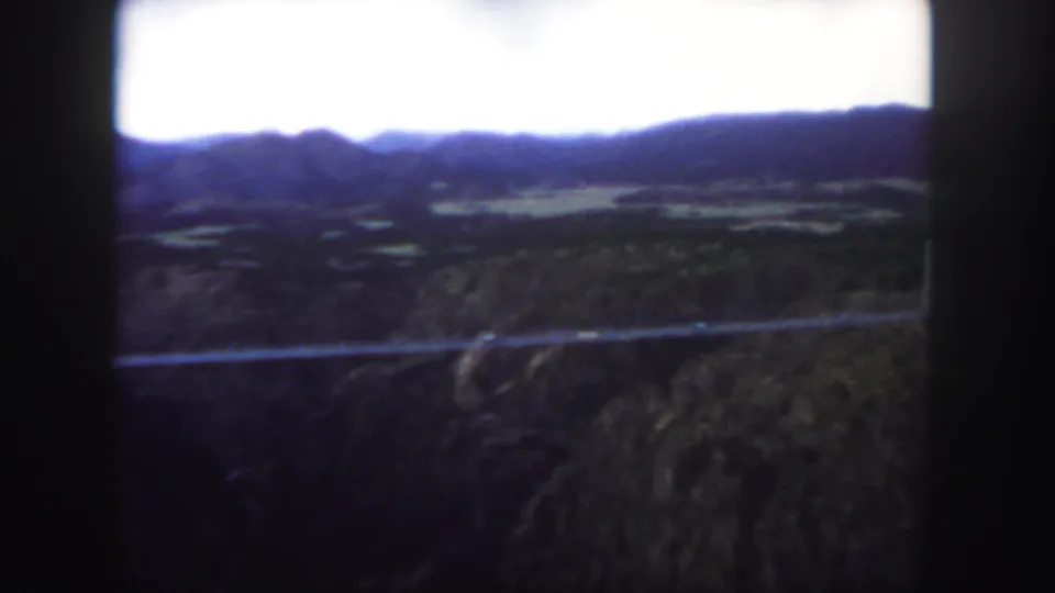 Colorado home-movie frame from 1958 showing the bridge as a thin line across rugged canyon terrain and distant hills.