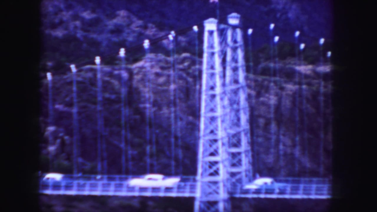 1958 Colorado 8mm frame showing cars on a suspension bridge beside steel towers and a rocky cliff backdrop.