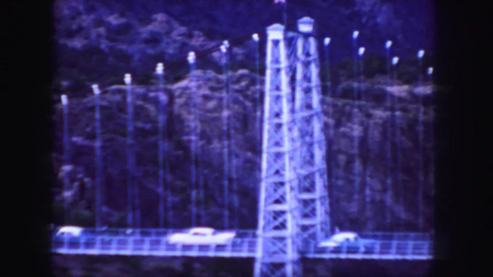1958 Colorado 8mm frame showing cars on a suspension bridge beside steel towers and a rocky cliff backdrop.