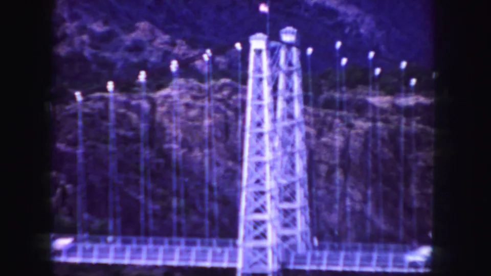 Blue-cast 1958 Colorado 8mm frame focused on a suspension bridge tower with cars partly visible on the span.