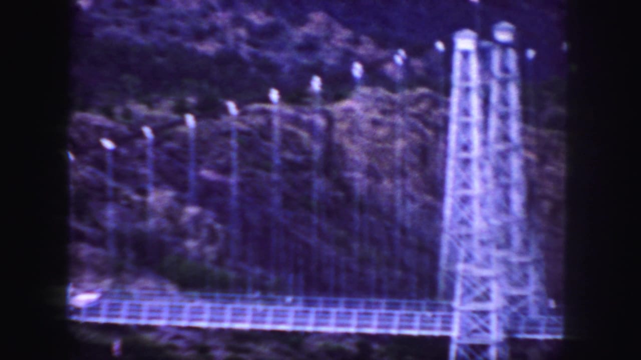 1958 Colorado 8mm frame showing a suspension bridge tower, bridge deck, poles, and rugged hillside vegetation.