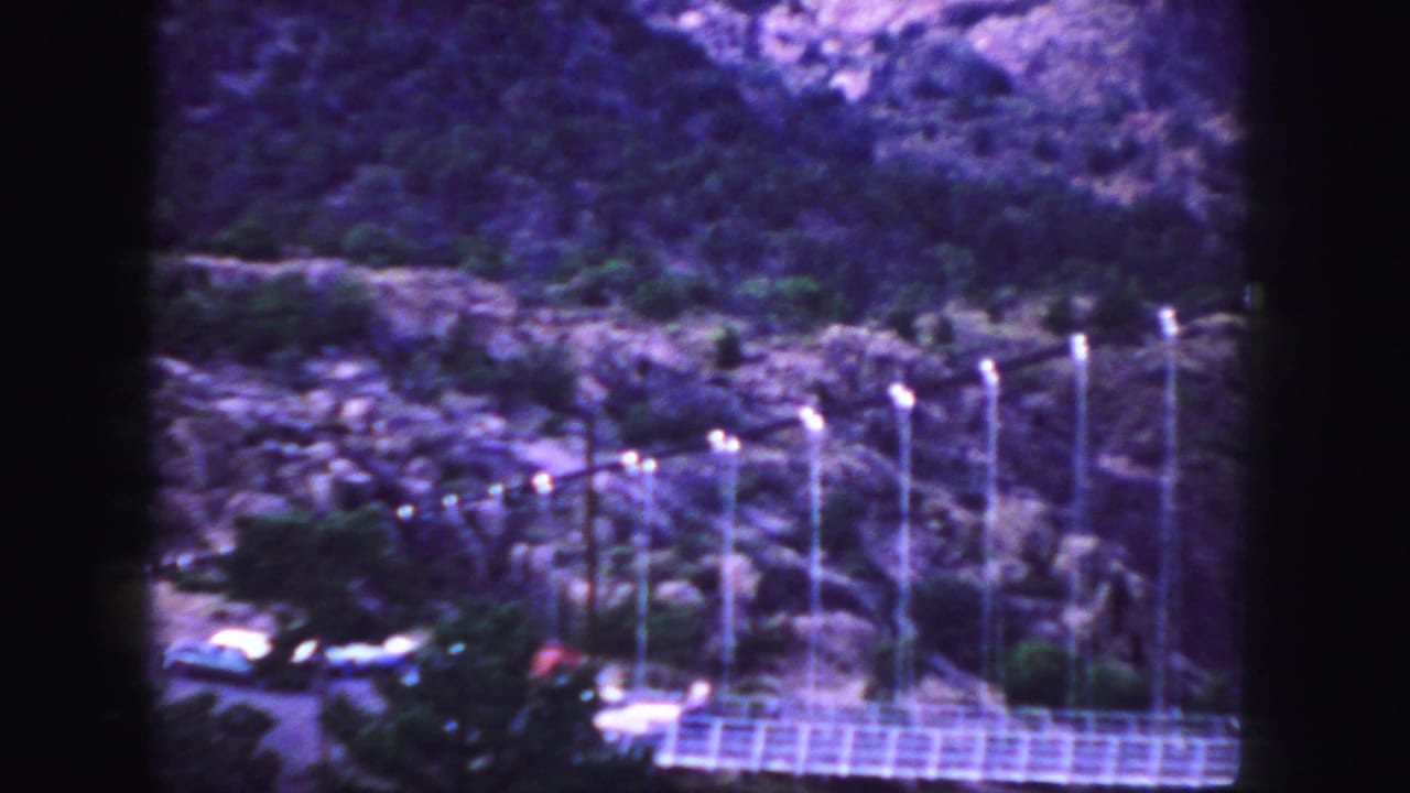 White car crossing a bridge below a rocky, tree-covered slope.