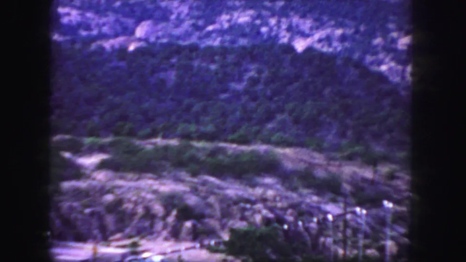 Colorado mountain scenery on 8mm film, with rocky terrain, low vegetation, a road, and partial bridge-area structures visible.