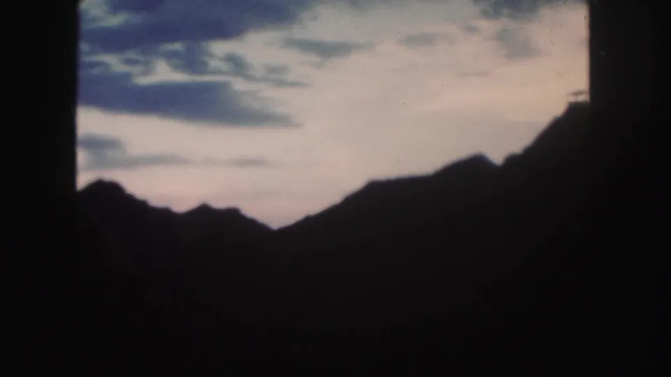 1958 Colorado home-movie frame with a dark mountain profile under a pale cloudy sky and strong black frame borders.
