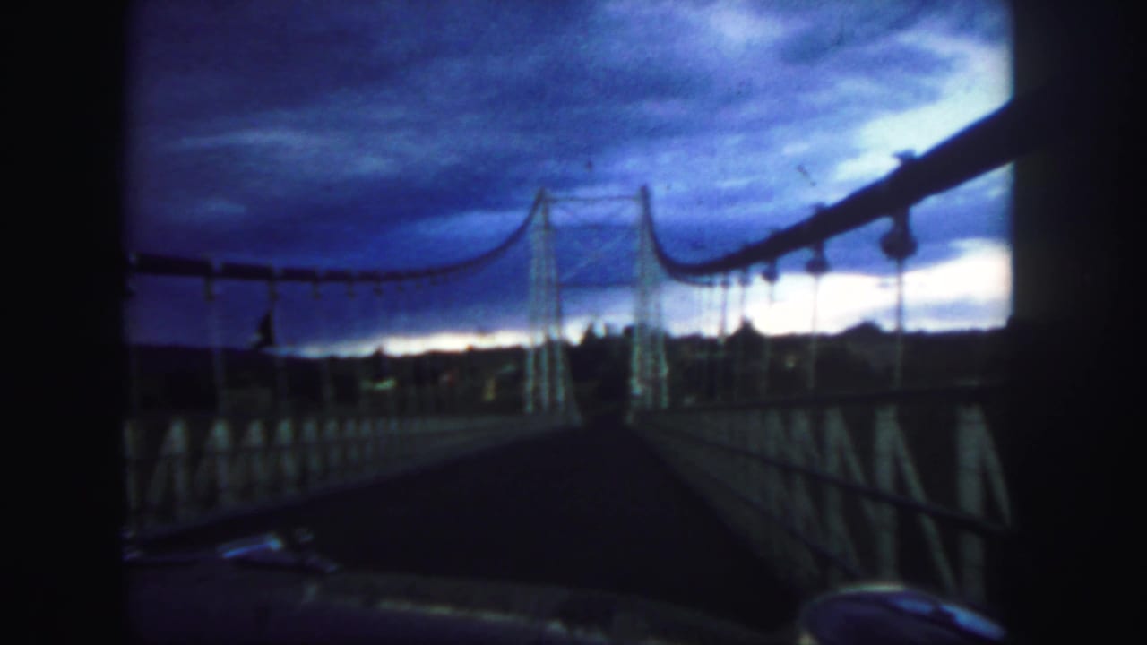 Vehicle approaches a suspension bridge beneath a dark cloudy sky.