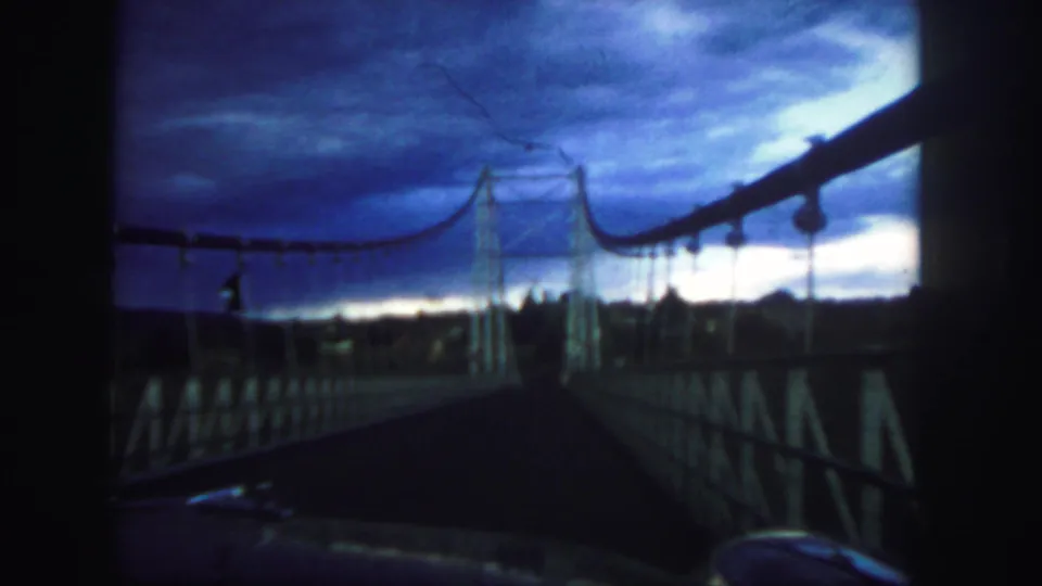 Color 8mm frame from Colorado in 1958 showing the bridge entrance, railings, and overhead cable line beneath a dramatic sky.