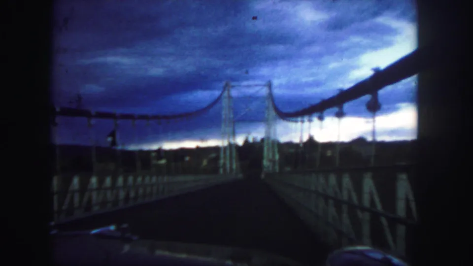 1958 Colorado 8mm frame showing a vehicle-perspective view along a suspension bridge with dark rails, cables, and storm-colored sky.
