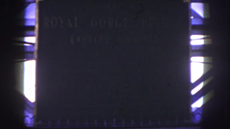 1958 home-movie frame of the Royal Gorge Bridge plaque, with faint lettering, a bright side opening, and visible 8mm grain.