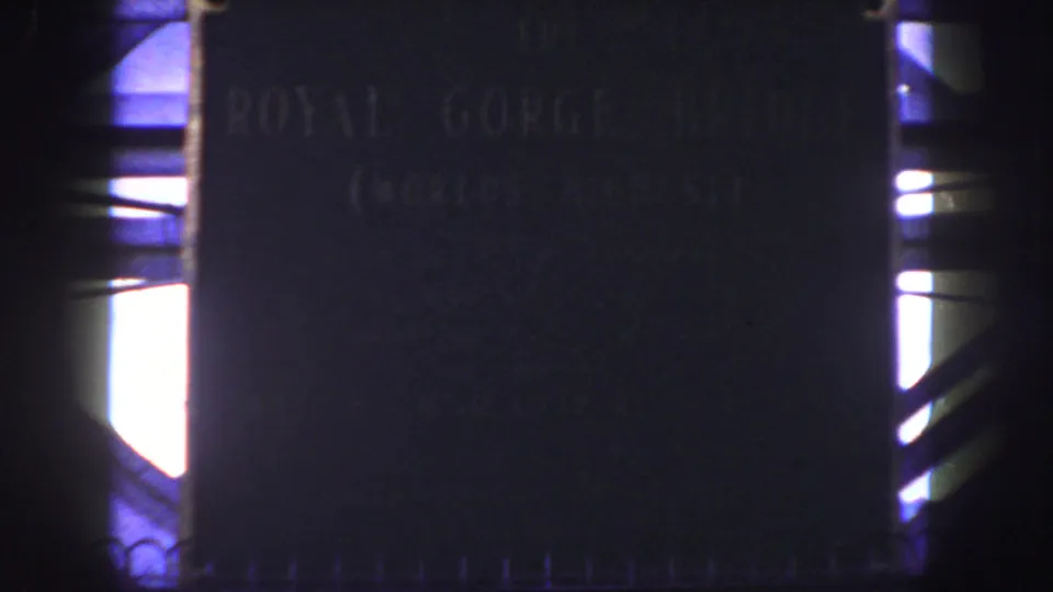 1958 home-movie frame of the Royal Gorge Bridge plaque, partially obscured by darkness and strong daylight contrast.