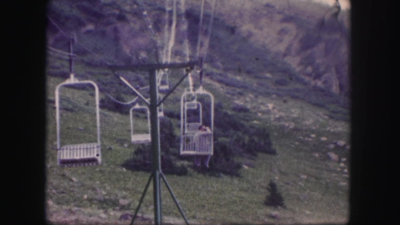 1958 Colorado 8mm frame showing white chairlift seats moving past a tower on grassy, rocky mountain terrain.