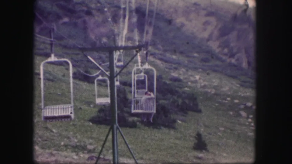 1958 Colorado 8mm frame showing white chairlift seats moving past a tower on grassy, rocky mountain terrain.