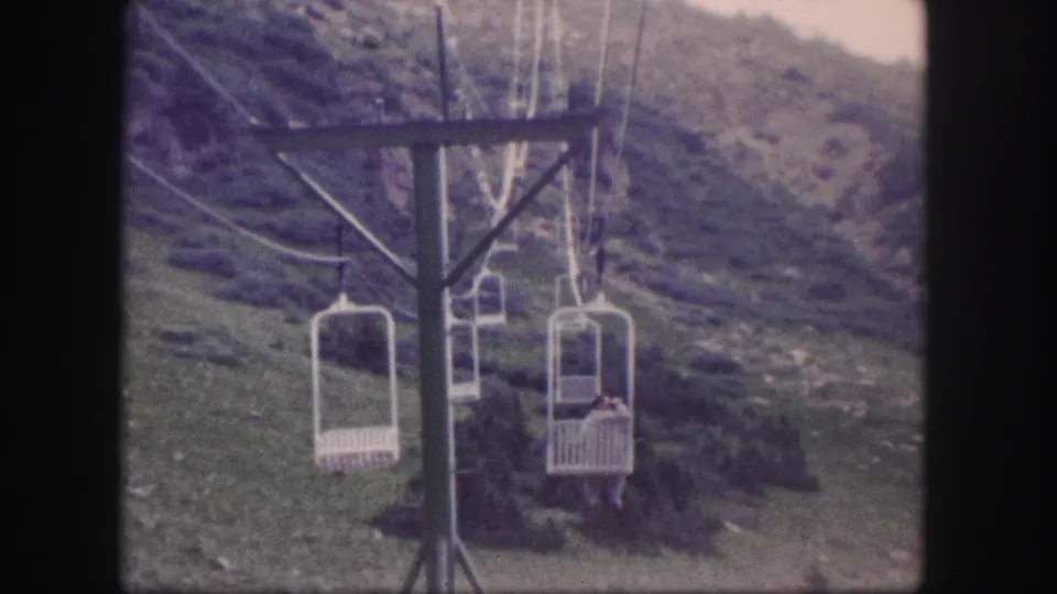 1958 Colorado home-movie frame of white chairlift seats and cables against a grassy, rocky mountain slope.