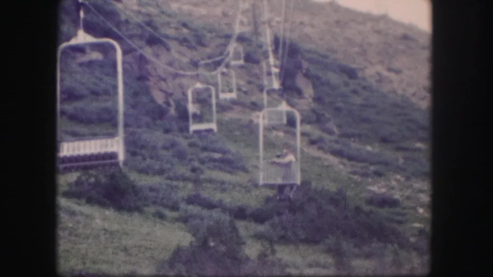 1958 Colorado 8mm frame of chairlift seats traveling over a green, rocky hillside with lift towers in the background.