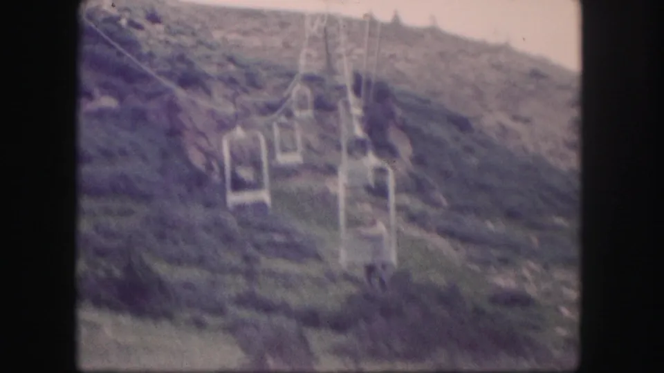 Soft 1958 Colorado 8mm frame showing multiple open chairlift seats suspended above a green mountain slope.