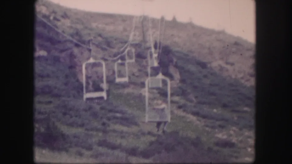 1958 Colorado 8mm frame showing a man seated on an open chairlift as it travels above a grassy mountain slope.