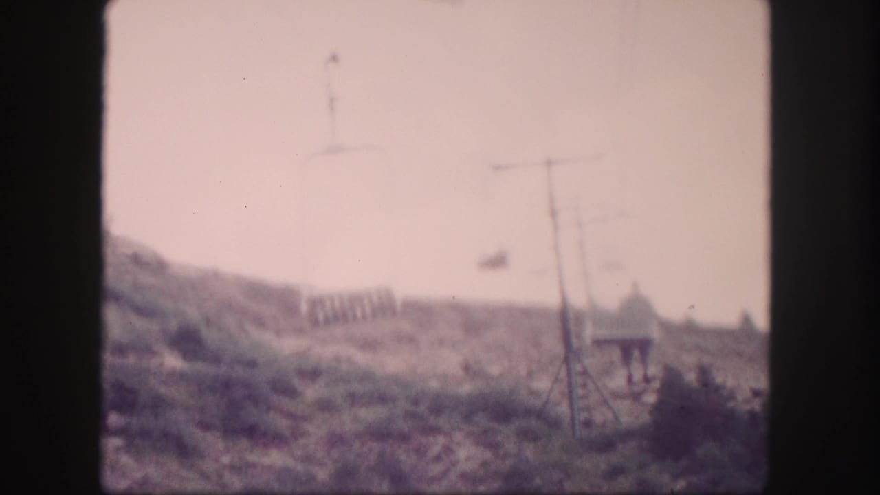 1958 Colorado 8mm frame with cable-car supports, lines, hillside scrub, a small structure, and a distant person near the slope.