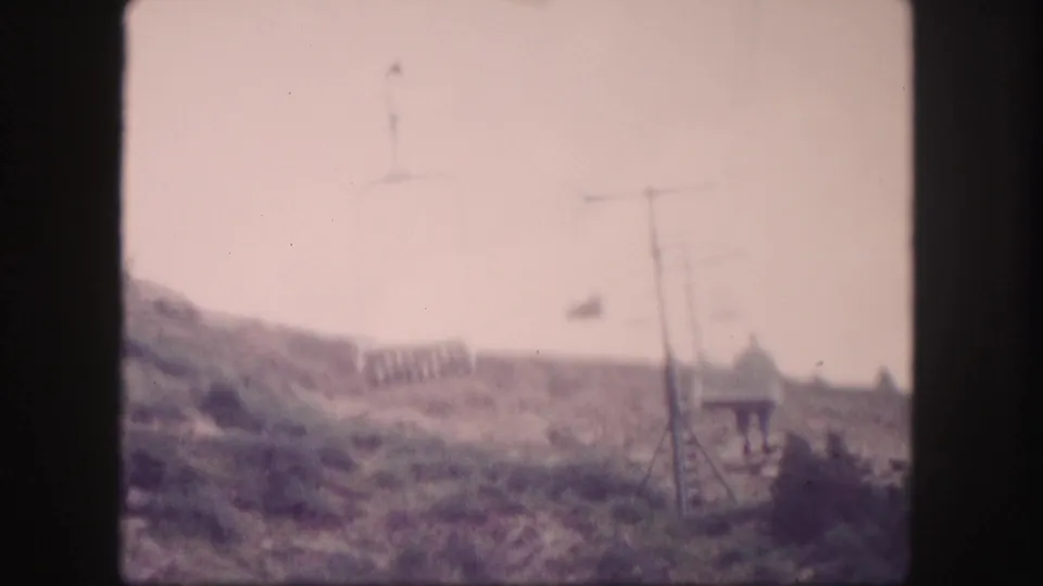 1958 Colorado 8mm frame with cable-car supports, lines, hillside scrub, a small structure, and a distant person near the slope.