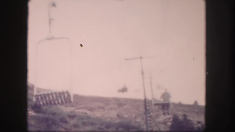 1958 Colorado 8mm frame showing cable poles, overhead lines, hillside vegetation, and a distant person by a small station-like structure.