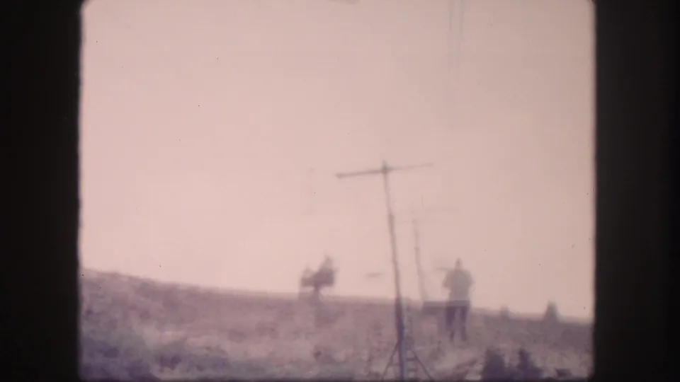 1958 Colorado 8mm frame with a distant standing figure, cable poles, overhead line, and open sunlit hillside.