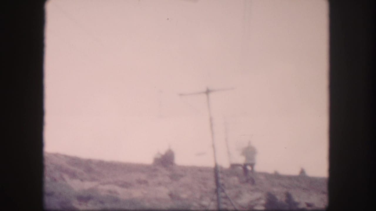 1958 Colorado 8mm frame showing distant chairlift riders moving along a mountain slope near a lift tower.
