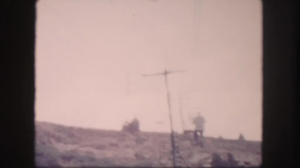 1958 Colorado 8mm frame showing distant chairlift riders moving along a mountain slope near a lift tower.