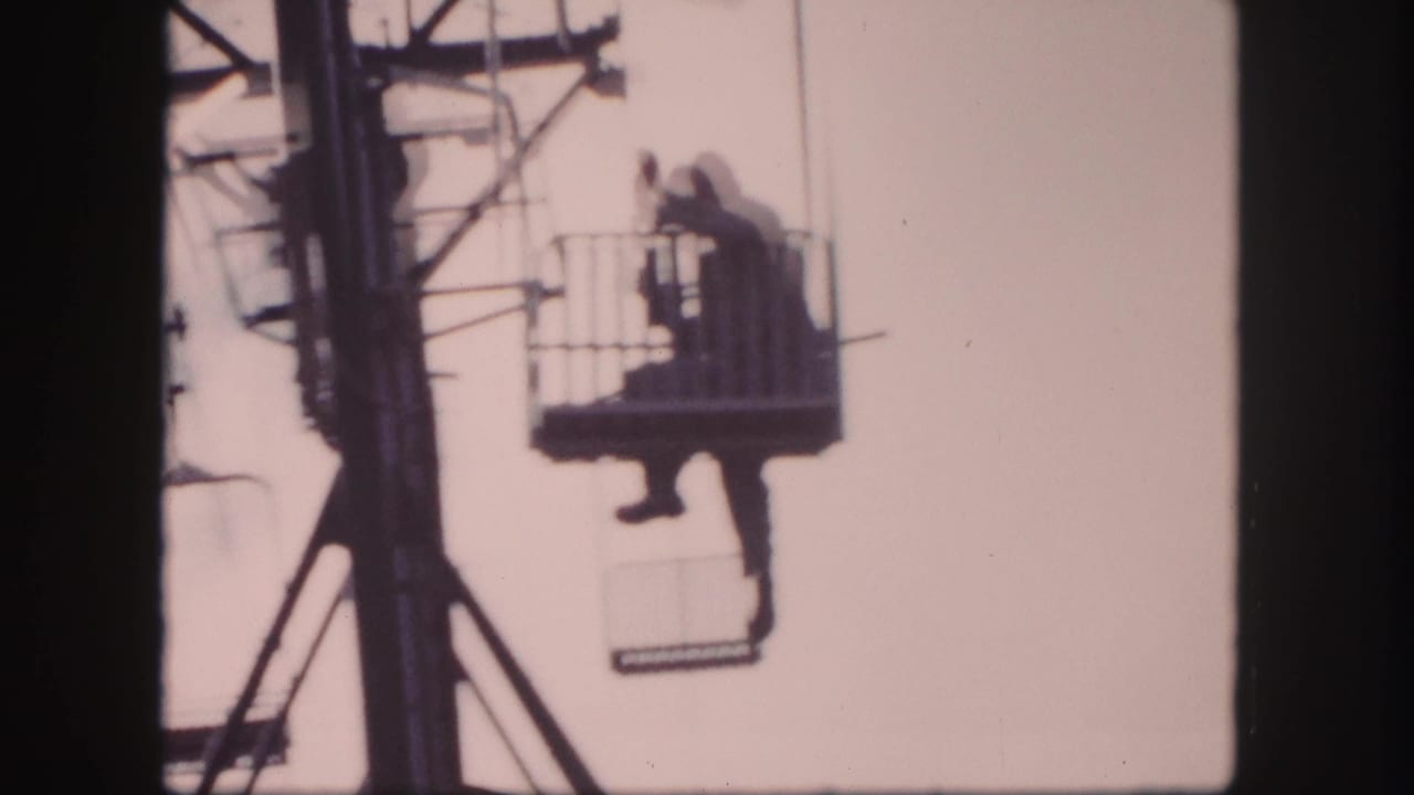 1958 Colorado home-movie frame showing a man in a dark coat seated in an open ski lift basket near a metal tower.