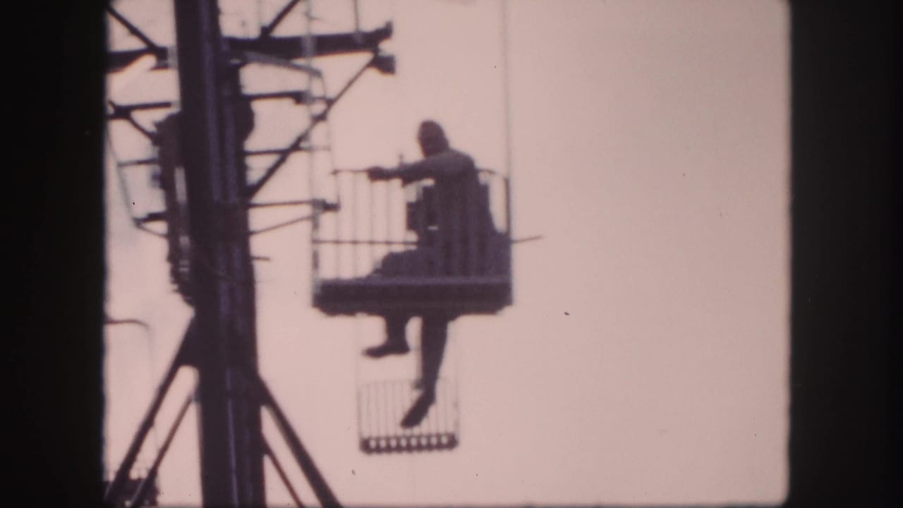 Man in a lift basket extends an arm while suspended beside the ski lift tower.