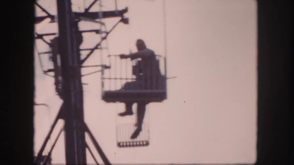 A man on a Colorado ski lift appears to wave from an open basket, silhouetted against a pale sky in 8mm footage.