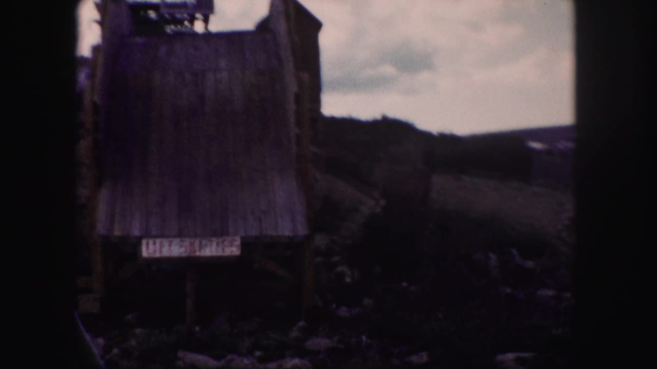 1958 Colorado 8mm frame showing a weathered wooden mountain structure with a small sign and distant slopes behind it.