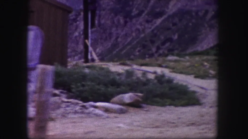 1958 Colorado frame showing a path, fencing, barrels, and a wooden structure in a rustic mountain lift-area setting.