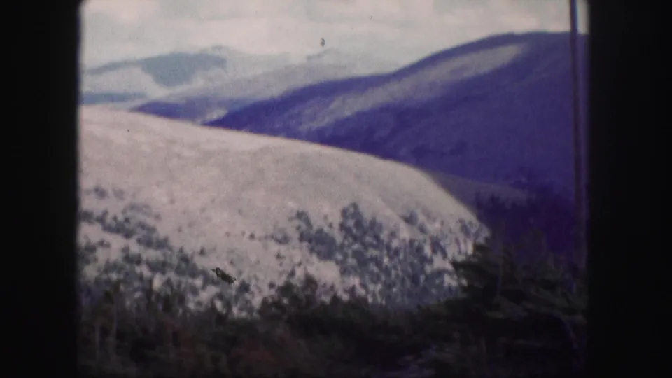1958 Colorado 8mm view across a pale mountain slope, distant ridges, cloudy sky, and dark evergreen foreground.