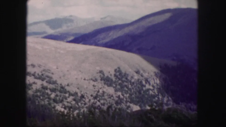 Soft 8mm frame from Colorado showing a pale mountain slope, blue-purple ridges, trees, and a cloudy sky.