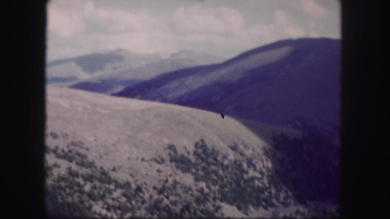 1958 Colorado 8mm frame showing layered mountain ridges, pale clouds, and dark wooded slopes with faded color and soft focus.