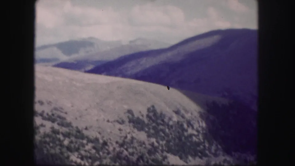 1958 Colorado 8mm frame showing layered mountain ridges, pale clouds, and dark wooded slopes with faded color and soft focus.