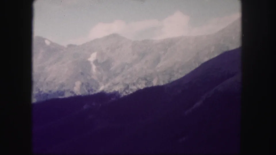 Vintage 1958 Colorado 8mm frame of distant mountain ridges, pale cloud cover, and a dark wooded slope in the foreground.