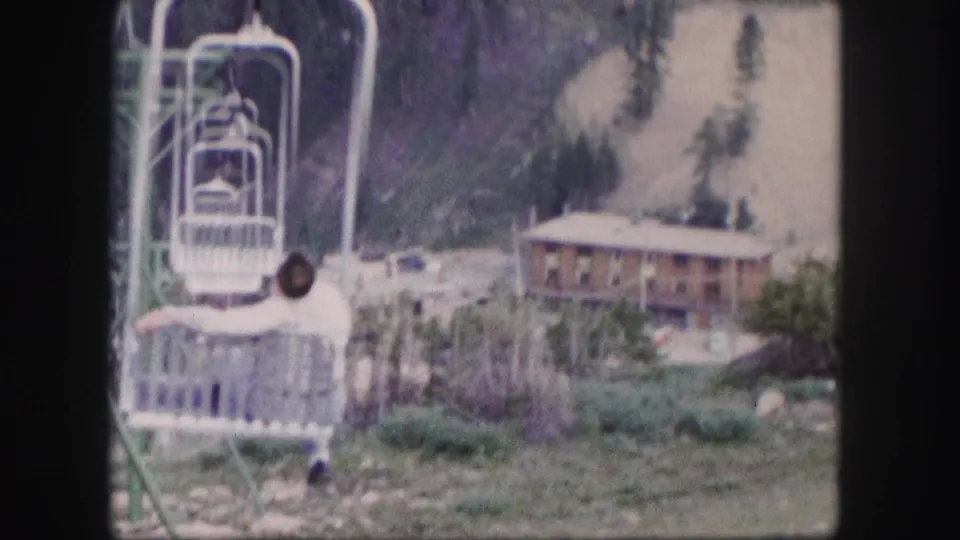 1958 Colorado tourist-center view with a man riding a cable car chair past trees and a rustic lodge.
