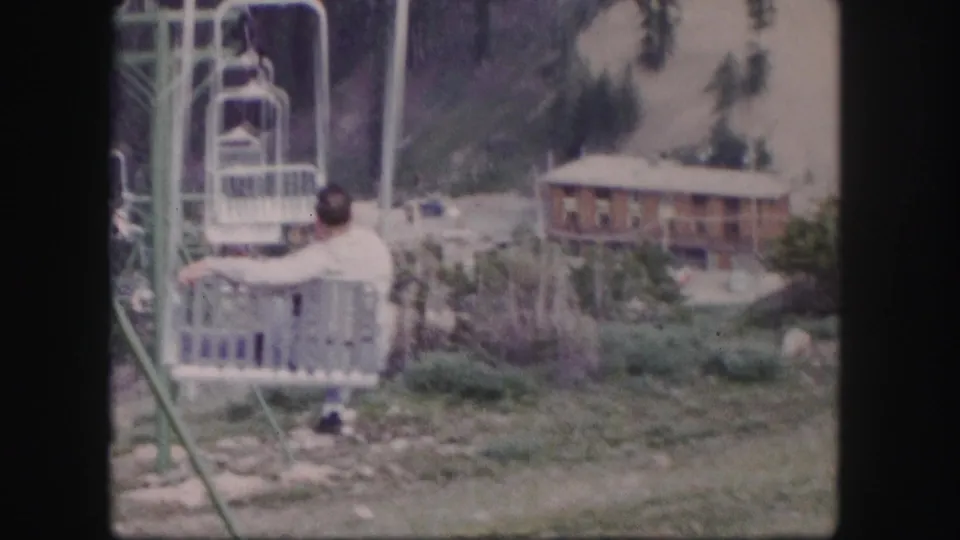 Man on a cable car chair in a Colorado mountain tourist area, with lodge, trees, and hillside behind him.
