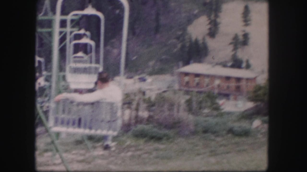 1958 Colorado 8mm frame showing a man on a ski lift near a rustic lodge and forested mountain slope.