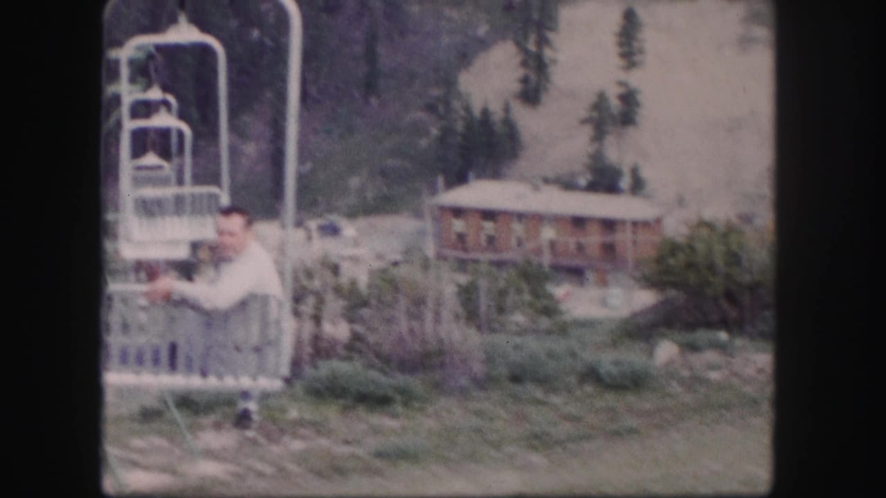 Man looks toward the camera while seated on a chairlift near a lodge.