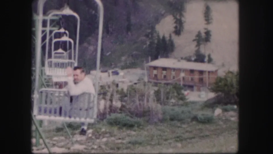 Colorado 1958 8mm frame showing a chairlift rider, metal lift structure, lodge, and wooded mountain background.