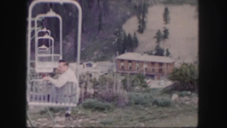 1958 Colorado resort frame with a man in a light shirt riding a chairlift beside a lodge and forested slope.
