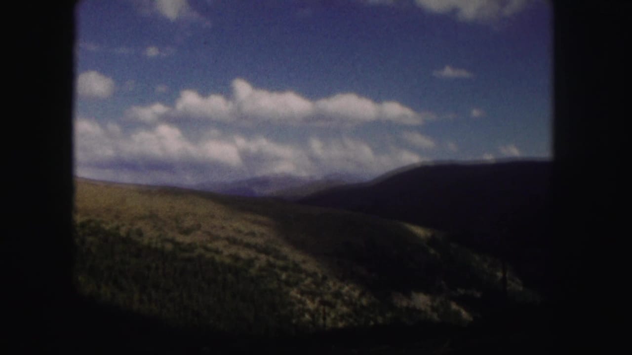 1958 Colorado 8mm frame showing rolling green hills, dark wooded slopes, distant ridges, and a cloud-filled blue sky.
