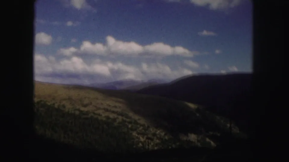 1958 Colorado 8mm frame showing rolling green hills, dark wooded slopes, distant ridges, and a cloud-filled blue sky.