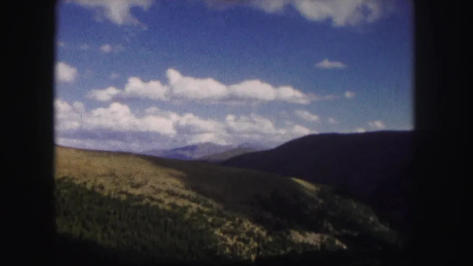 1958 Colorado scenic frame with a green foreground hillside, dark ridge line, distant peak, and scattered clouds in blue sky.