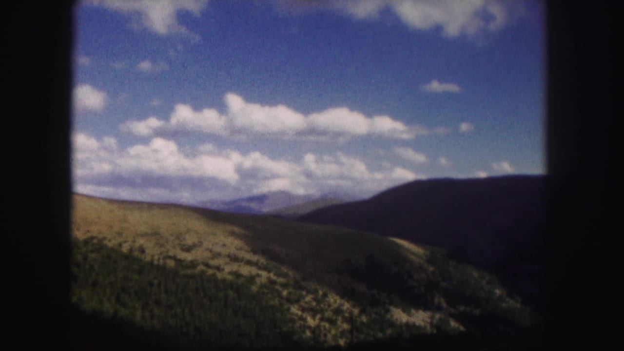 1958 Colorado 8mm view of green rolling hills, darker forested slopes, distant ridges, and scattered clouds in a blue sky.