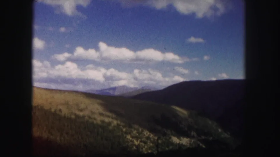 Colorado home-movie frame showing green hills, a shadowed mountain ridge, distant peaks, and bright clouds across a blue sky.