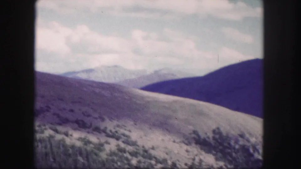 1958 Colorado landscape frame with a pale grassy slope in front of layered blue mountain ridges and a soft cloudy sky.