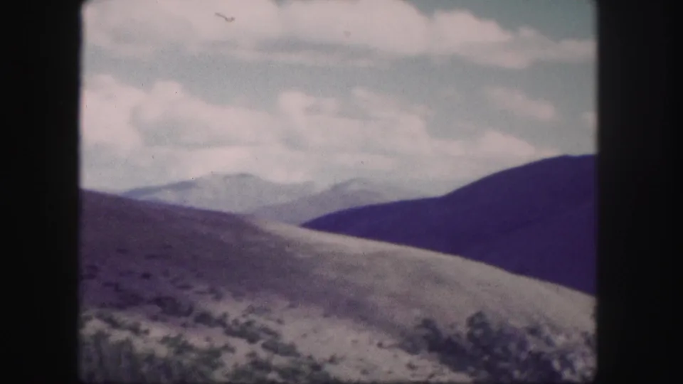 Colorado 8mm frame of an open hillside, darker mountain ridge, distant peaks, and muted clouds with visible film softness.