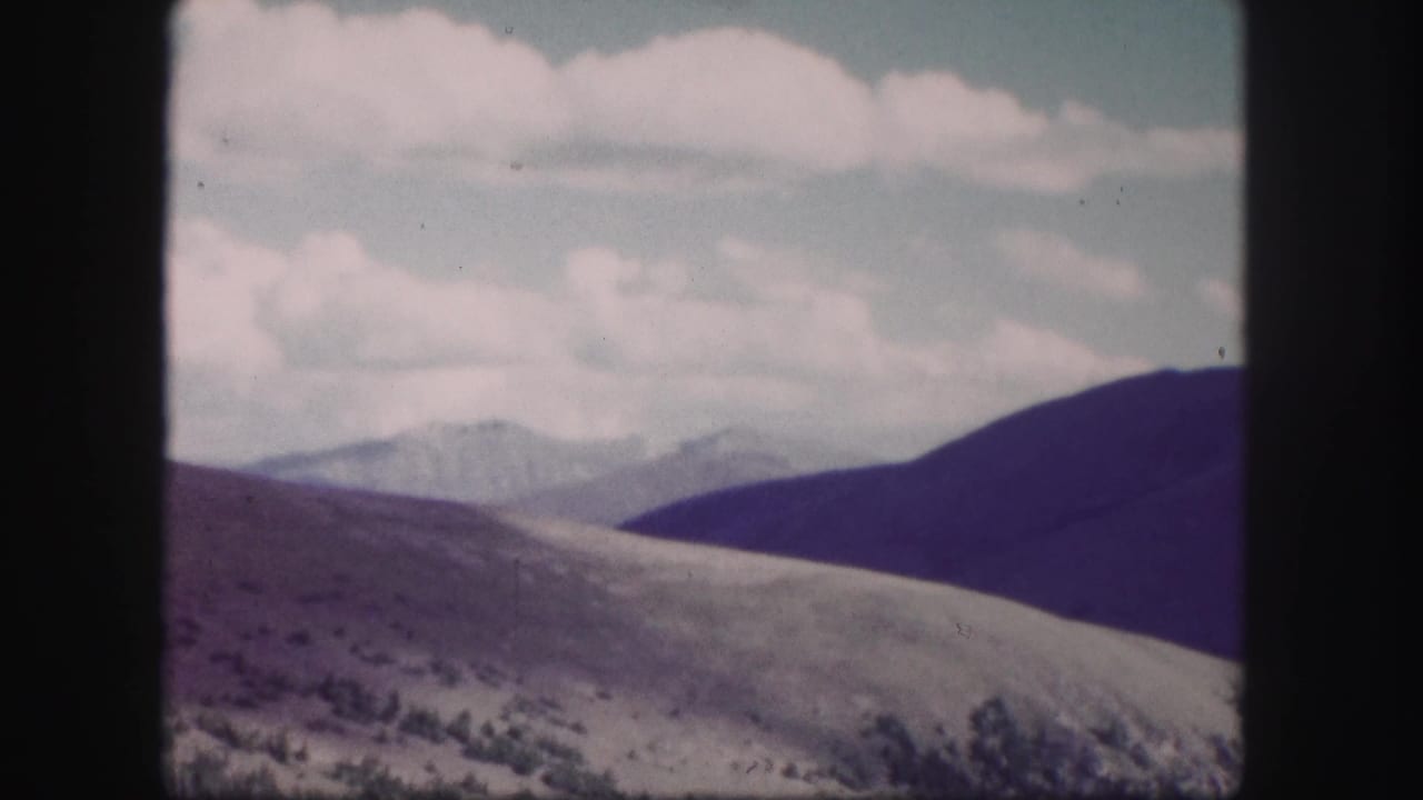 1958 Colorado 8mm frame showing open rolling hills, distant mountain ridges, sparse vegetation, and pale clouds overhead.